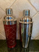 Photo showing two cocktail bar shakers side by side on a countertop. Left is red glass with stainless steel top and crisscross pattern, right is stainless steel with branding.