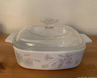 Vintage CorningWare small white square casserole dish with pastel floral design and clear glass lid with knob, placed on wooden surface.