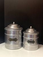 Photo showing the pair of vintage aluminum canisters with lids, stacked side-by-side against a black background. Larger canister labeled 'FOUR'.