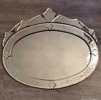 Front view of the vintage ornate oval Venetian beveled glass wall mirror laid flat on floor showing detailed etched floral and leaf design on the frame glass.