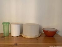 Four vintage Tupperware items arranged side by side including a green drink holder, white canister, beige cake holder, and orange bowl.