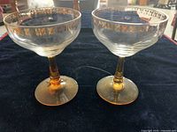 Pair of vintage champagne glasses with amber stems and gold leaf designs on rims placed on a dark cloth.