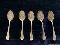 Five silver plated small spoons arranged horizontally on a dark cloth background, showing general condition and design shape.