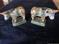 Pair of ceramic cow figurines side by side on a dark cloth surface, showing pattern and colors.