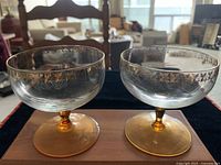 Two crystal champagne glasses with clear bowls, gold leaf pattern on the rim, amber stem and base, placed on wooden surface.