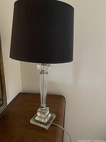Table lamp with clear glass base and gray fabric cylindrical shade sitting on a wooden surface, plugged in with a visible power cord.