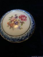 Close up top view of circular lid of small trinket dish showing detailed floral pattern with pink rose, purple and orange flowers, surrounded by blue border with gold trim.
