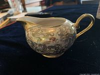 Side view of a bone china creamer with gold detailing and black floral pattern sitting on a dark cloth