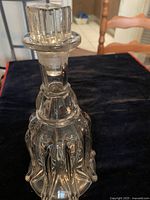 Photo of a clear glass vintage-style decanter with intricate design on the body and a faceted stopper on top, placed on a dark cloth with a chair in the background.