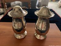 Pair of vintage silver plated salt and pepper shakers on wooden surface with black furniture in background. Both items have a reflective body and decorative scalloped tops with perforations for dispensing.