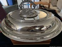 Top and side view of the silver-plated serving dish showing the oval shape, curved lid with floral motif base for handle, beaded loop handle, and raised beading around rim.