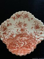 Close-up of scalloped saucer showing red floral transferware clover pattern with gold trim on white porcelain.