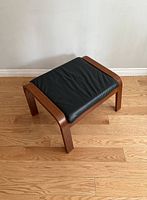 Front angled view of the vintage IKEA POÄNG footstool showing the curved wooden legs and black cushion.