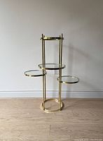 Front view of vintage brass and glass multi-tier plant stand against a white wall, displaying four round glass shelves of varying heights.