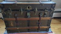 Front view of antique steamer trunk showing wood panels, metal corners, leather straps, and central lock with latches.