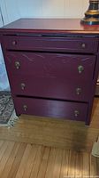Front view of burgundy wooden chest showing three drawers with brass handles and carved wood detail in middle drawer.