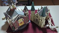 Front and angled view showing two ceramic Christmas village buildings, one red brick with a clock face and the other a church with teal spires, both with power cords.