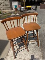 Two wooden counter stools with spindle backrests and curved top rails placed outdoors in sunlight.