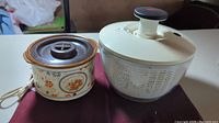 Photo of electric potpourri crock with floral design next to white plastic salad spinner with pump lid on a fabric surface.