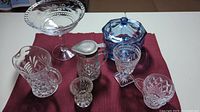 Full group of seven pieces on a burgundy placemat showing blue lidded sugar bowl, clear pitcher, candy dish, cup, bowl, and two small glass pieces.