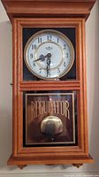 Front view of wooden regulator wall clock with glass front showcasing white dial with black numbers and decorative hands, brass pendulum, and word 'REGULATOR' on glass.