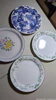 Four dinner plates showing different floral and grape-themed designs placed side by side on a surface