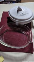 Clear glass oval casserole dish with handles and white ceramic oval casserole dish with embossed leaf lid on red cloth surface