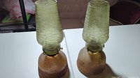 Two green textured glass oil lamps side by side with visible wick adjustment metal features on bases and matching textured glass chimneys.