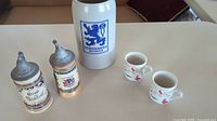 Photo showing two lidded ceramic beer steins, one large Löwenbräu Munich ceramic mug, and two small white decorative mugs placed on a table.