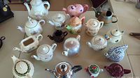 Full view of 18 assorted teapots arranged on a table showing variety in design and materials including porcelain and metal