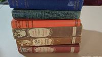 Six vintage hardcover books by Louisa May Alcott stacked horizontally showing spines and some covers. Colors vary from blue, green, orange, brown, maroon. Wear on covers and spines visible.