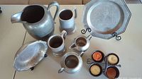 Vintage pewter tableware including pitcher, mugs, cream and sugar pots, and plates, alongside four citrus-scented candles in black holders.