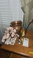 Photo showing hammered copper bucket, brass eyeglass holder with back scratcher inside, and cherry blossom tree on marble base placed on wooden table.