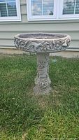 Full view of bird bath standing on grass against house siding, showing the floral and hummingbird relief patterns around the basin and pedestal.