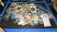 Overview of multiple collectible pins on black surface, mostly sealed in plastic bags, including badges and event pins.