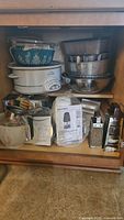 Kitchen cabinet shelf filled with Rival crock pot, nested mixing bowls with lids, metal graters, cake pans, Pyrex bowls with blue floral pattern, and other kitchen utensils including wrap dispensers and measuring cups.