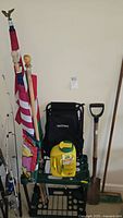 Photo showing multiple flags on poles, a black filled backpack on a green garden tool holder cart with a jug of plant food in front, a broom and shovel propped against the wall, and fishing rods visible to the left.