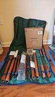 Green fabric carrying case containing a set of BBQ tools with wooden handles including spatula, fork, tongs, brushes, and the boxed grill cover.
