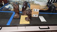 Three carved dark brown ironwood owls displayed on a surface, each with distinct carving details. Two cardboard boxes beside them likely containing the pot belly owl figures. Blue tape and miscellaneous items visible in background.