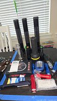 Multiple assorted flashlights displayed on table including 3 black Maglite flashlights, silver tactical flashlight, blue LED flashlight, red flashlight, batteries, and accessories in plastic bag.