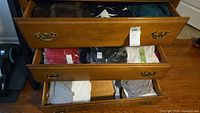 Three wooden drawers open showing assorted men's clothes including sweaters, t-shirts, and shirts, mostly size XXL.