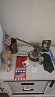 Overall view of military memorabilia collection on a white cabinet, includes the handmade metal cannon, American flag, patches, sealed iron-on patch, small box, brass-colored door knocker, and small light device.