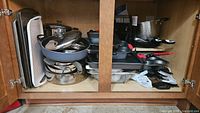 Kitchen cabinet with pots, pans, lids stacked and various kitchen tools inside.