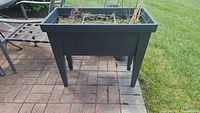 Front view of the black plastic raised garden bed showing soil and dried plants inside, and part of the surrounding patio and grass.