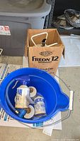 Lot showing Dupont Freon 12 cardboard box, blue plastic bucket containing two Arctic Air 134-A refrigerant cans, and white hook tool.