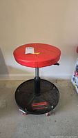 Front view of US General pneumatic adjustable roller seat stool showing red round seat, black metal base, and casters on floor.