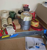 View showing assortment of string, twine, and various adhesive tapes including duct tape, packing tape, and brand labeled tapes with a tape dispenser