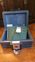 Open strong box with three green coin books inside and key attached to lock.
