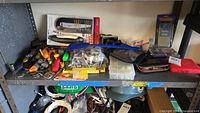 Wide view of a shelf holding multiple assorted tools and kits including screwdrivers, cutters, tape measure, wrench, electric stud finder, nails kits, trowels, and a blue flexible tool holder.