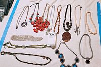 Overhead view of assorted necklaces laid out on white surface, featuring various styles such as beaded, metal chain, and pendant necklaces in multiple colors including coral red, black, silver, and blue accents.